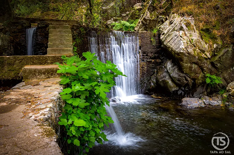 Cascata junto ao antigo açude na Praia Fluvial de Nossa Senhora da Piedade, rodeada por vegetação e rochas da serra da Lousã.