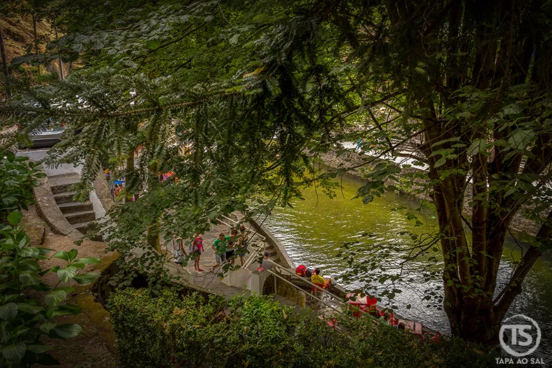 Vista parcial da Praia Fluvial de Nossa Senhora da Piedade através das árvores, com visitantes junto à margem e água verde esmeralda da ribeira.