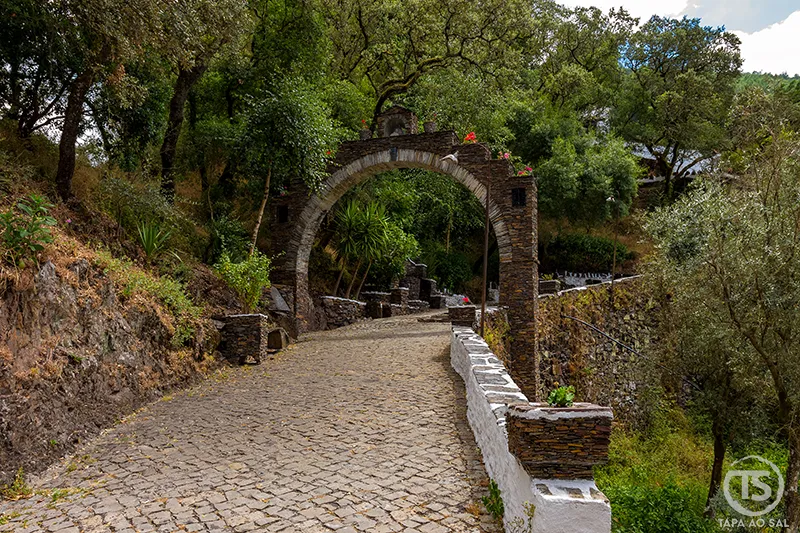 Arco em pedra na entrada do Santuário de Nossa Senhora da Piedade, rodeado por vegetação e calçada tradicional, na serra da Lousã