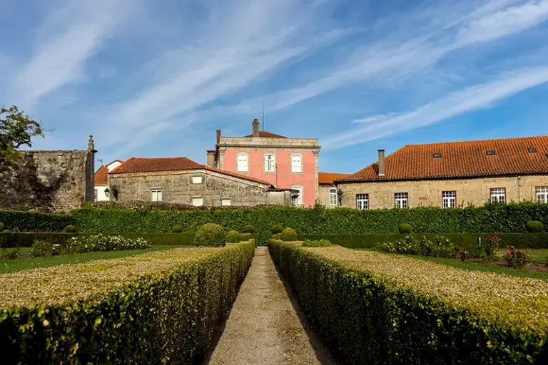 Jardim formal com sebes geométricas e edifício senhorial no centro de Santar Vila Jardim, em dia claro com céu azul.