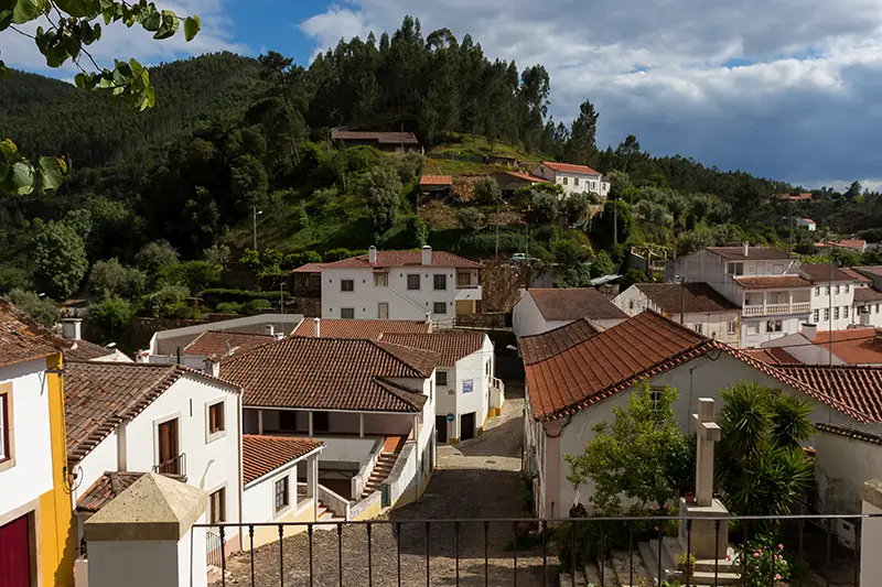 Vista panorâmica das casas de Dornes distribuídas pela encosta, com telhados de telha vermelha e o Rio Zêzere ao fundo, envoltos por vegetação verdejante.