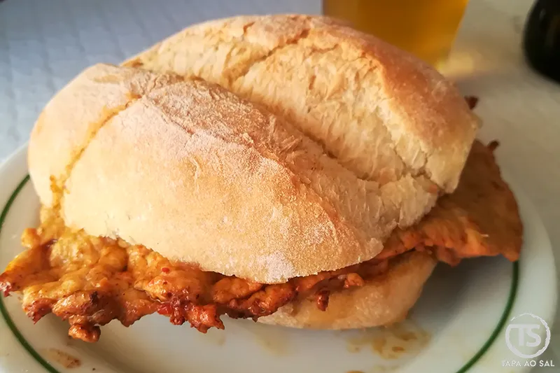 Bifana portuguesa tradicional servida em pão crocante, com carne marinada e cerveja ao fundo.