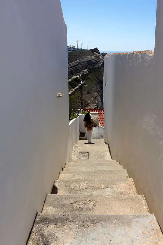 Mulher a descer escadaria estreita entre paredes brancas nas Azenhas do Mar, em Sintra.