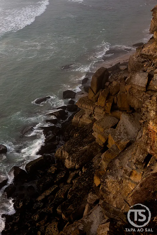 Falésia rochosa das Azenhas do Mar com o mar a bater nas pedras ao entardecer.