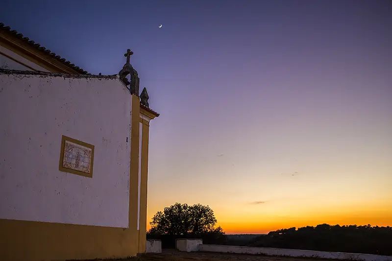 Ermida da Senhora da Graça em Nisa ao pôr do sol, com céu violeta e crescente lunar no horizonte.