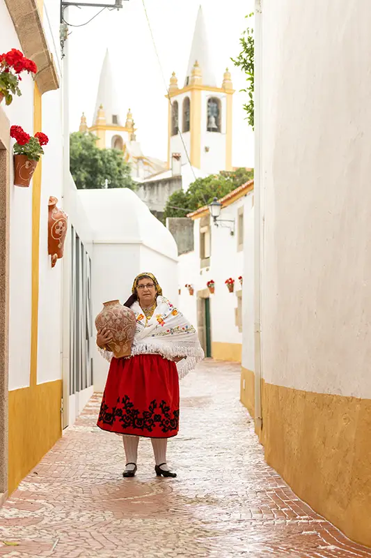 Mulher em traje tradicional de Nisa com uma bilha de barro, numa rua típica decorada com vasos de flores e vista para as torres da Igreja Matriz ao fundo.