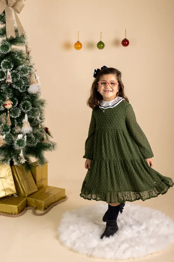 Criança sorridente em sessão de Natal, sentada junto à árvore decorada e presentes