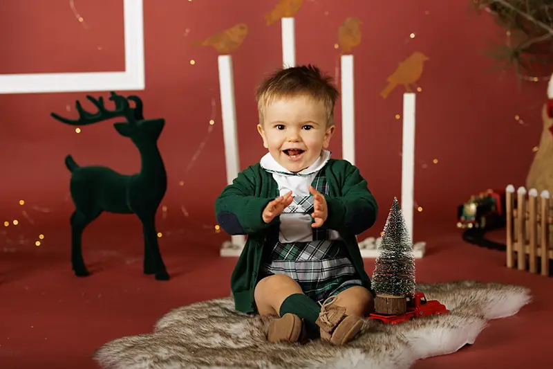 Criança sorridente em sessão fotográfica de Natal, sentada em cenário festivo com rena e árvore decorativa.