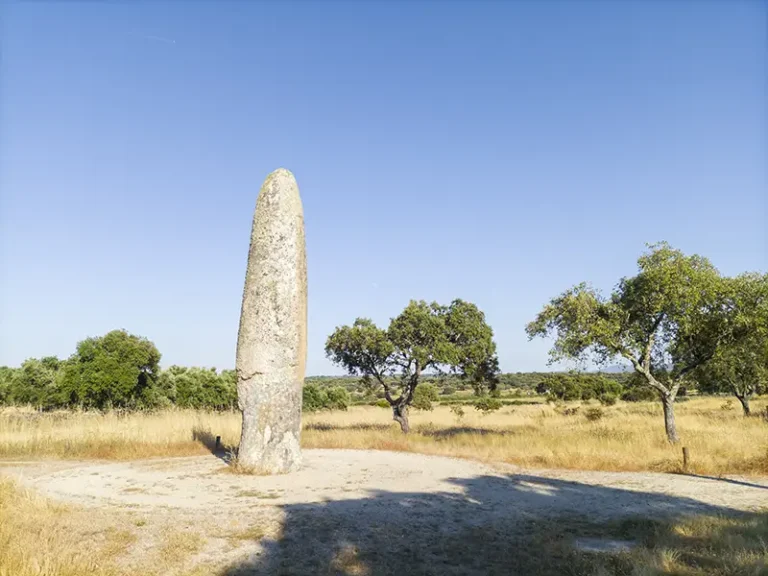 Menir da Meada ao entardecer, rodeado por paisagem natural alentejana com azinheiras e céu limpo