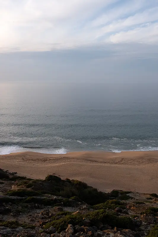 Praia deserta do litoral atlântico português vista do alto da falésia, com mar calmo e luz suave — exemplo de paisagem costeira que não podes perder ao explorar o que visitar em Portugal.