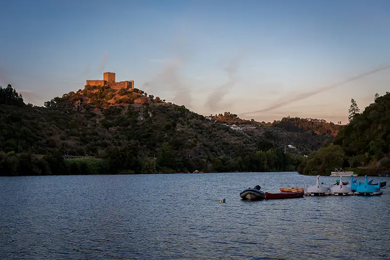 Castelo de Belver iluminado pela luz dourada do entardecer, visto desde o Tejo com pequenos barcos ancorados — cenário histórico e romântico a considerar quando decides o que visitar em Portugal.