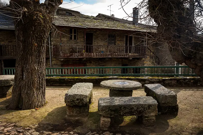Mesa e bancos de granito sob árvores despidas na aldeia comunitária de Rio de Onor, Trás-os-Montes — paragem autêntica a incluir quando procuras o que visitar em Portugal num roteiro rápido pelo Norte.