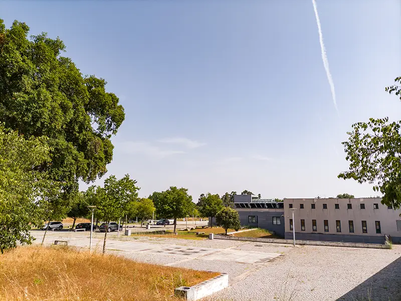 Estacionamento e edifício das Termas da Fadagosa de Nisa, rodeados de árvores e céu limpo num dia de verão.