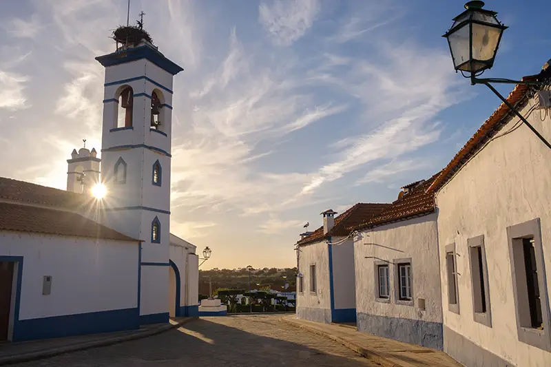 Aldeia de Santa Susana ao pôr do sol, com casas caiadas e torre da igreja iluminada pela luz dourada do verão — cenário típico para quem procura o que visitar em Portugal nesta estação.