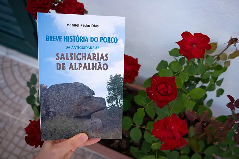 Livro “Breve História do Porco da Antiguidade às Salsicharias de Alpalhão”, de Manuel Pedro Dias, segurado por uma mão junto a um vaso com rosas vermelhas