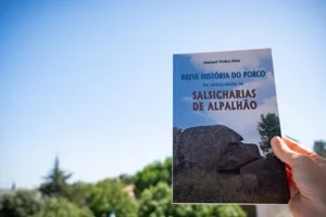 Capa do livro Breve História do Porco da Antiguidade às Salsicharias de Alpalhão, segurado por uma mão ao ar livre com céu azul e vegetação ao fundo