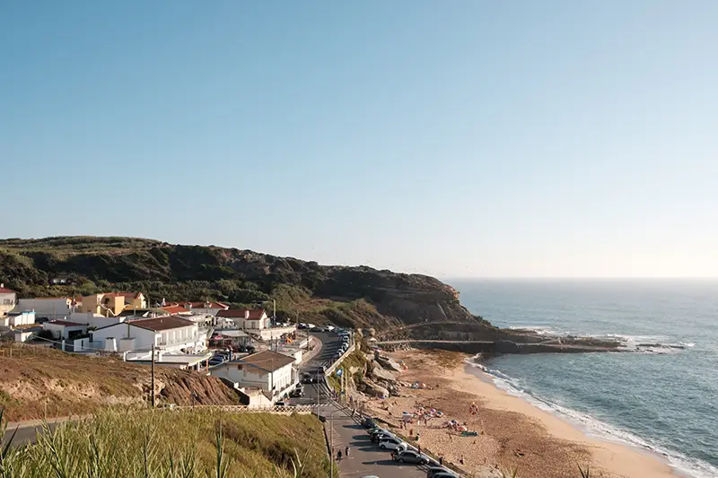 Vista panorâmica da Praia de Porto Dinheiro com zona de estacionamento, casas junto ao mar e o areal a estender-se até ao oceano Atlântico.
