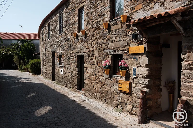 Fachada em pedra da Casa do Tear na aldeia da Figueira, com vasos floridos e detalhes tradicionais.