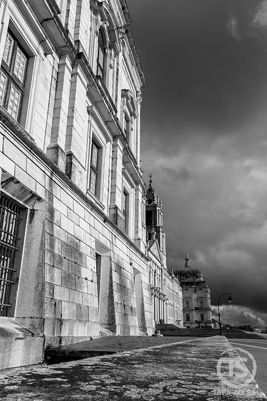 Detalhe lateral do Palácio Nacional de Mafra sob céu nublado em fotografia a preto e branco.