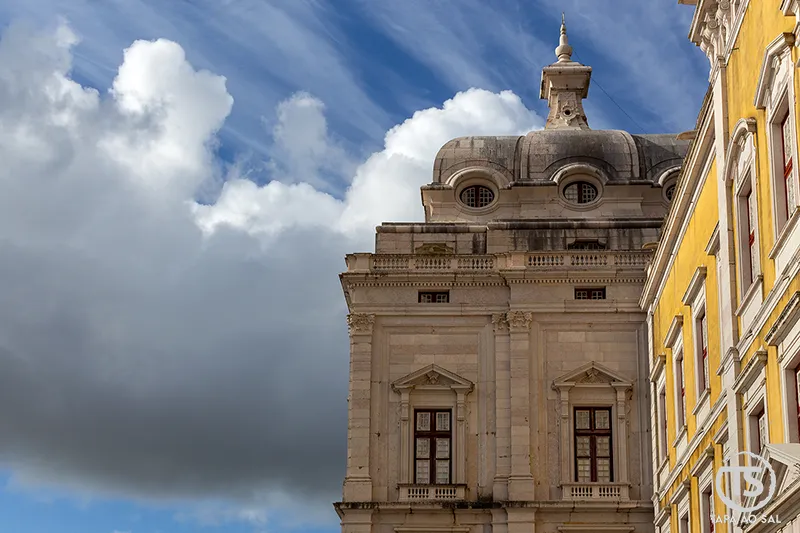 Detalhe arquitetónico do Palácio Nacional de Mafra com fachada amarela e cúpula barroca contra o céu nublado