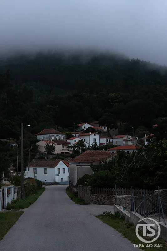 Estrada a entrar numa aldeia serrana envolta em nevoeiro, evocando alugar carro em Portugal para explorar o interior