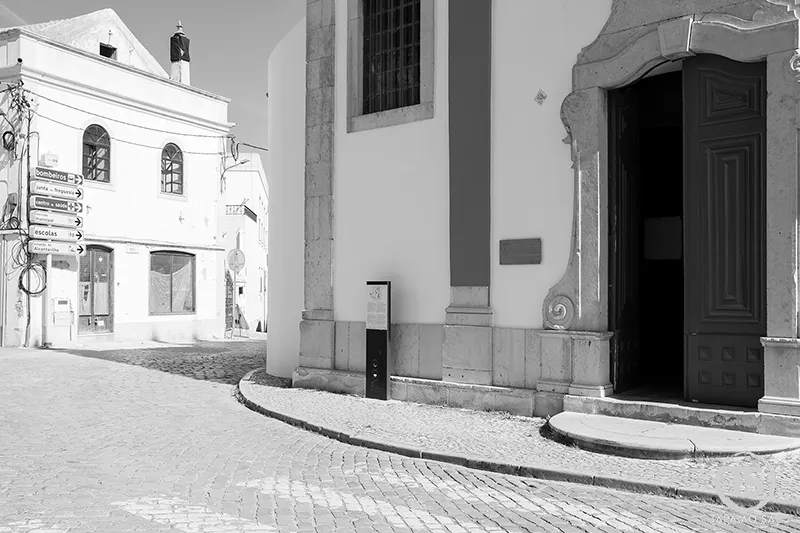 Rua curva junto à igreja matriz de Alcantarilha com fachadas brancas e porta aberta