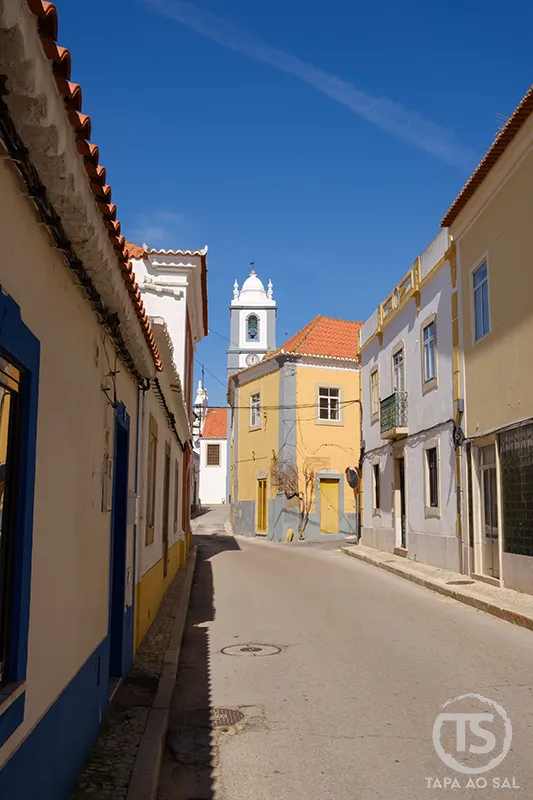 Rua de Alcantarilha com casas coloridas e torre da igreja ao fundo