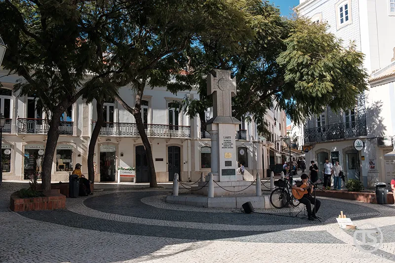 Praça no centro histórico de Lagos com monumento, calçada portuguesa e músico de rua