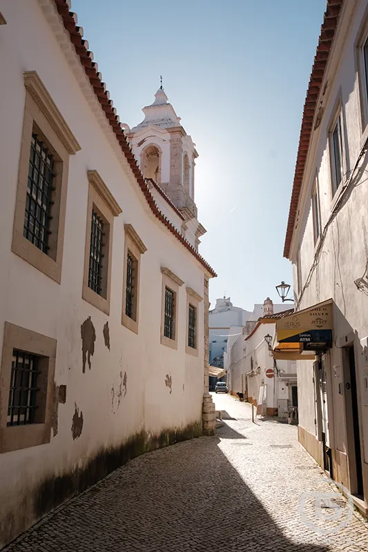 Rua do centro histórico de Lagos com calçada portuguesa e torre de igreja ao fundo